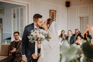 Bride and groom share first kiss at wedding ceremony while guests celebrate in elegant white venue.