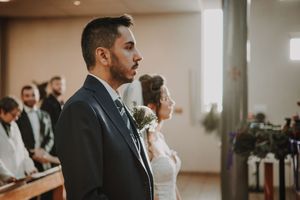 Groom in navy suit stands at church altar beside bride in white dress during wedding ceremony.