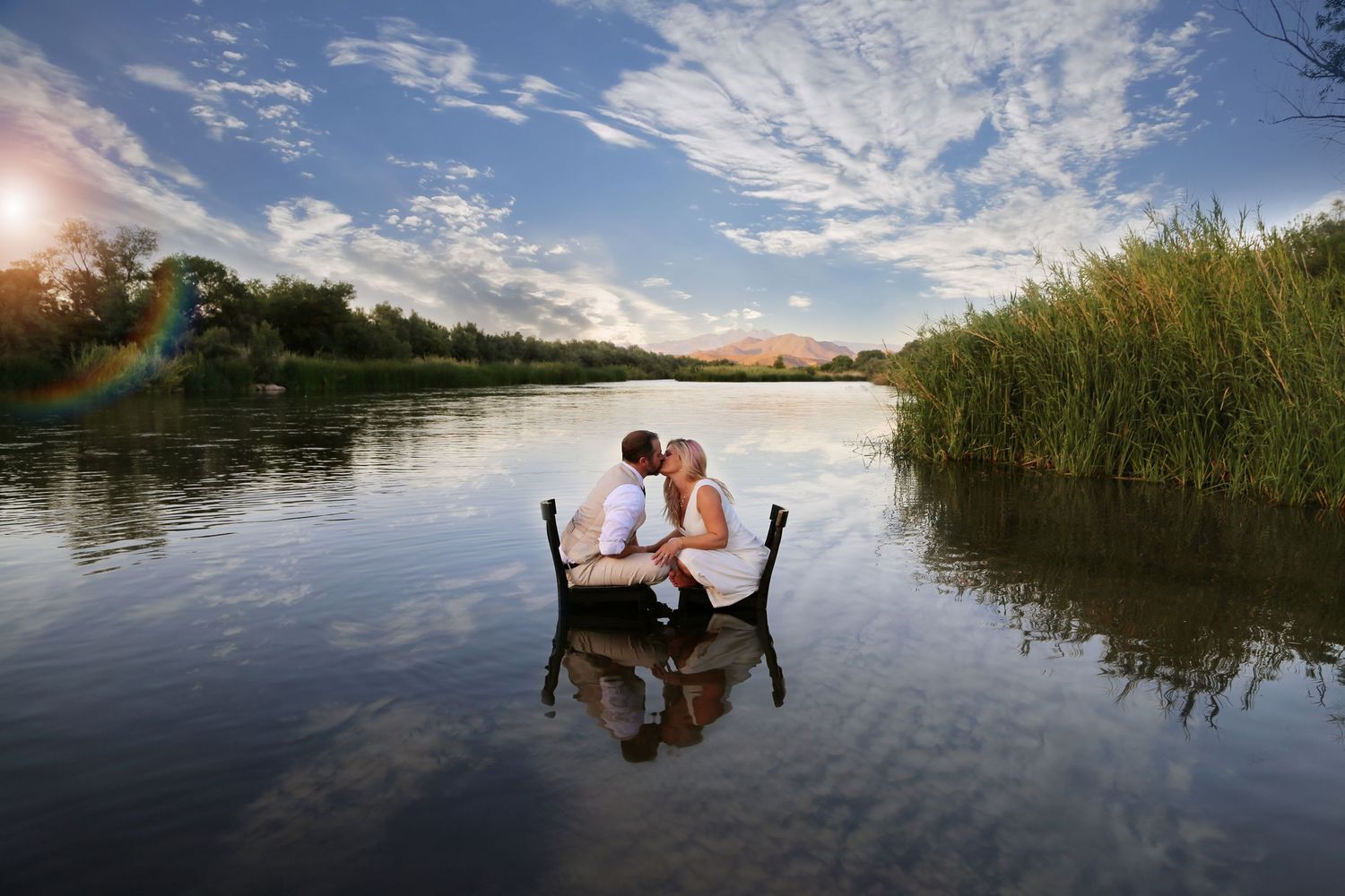 Couple shares a kiss while seated on a small boat in a calm lake, with reeds, trees and a lens flare under a bright blue sky.