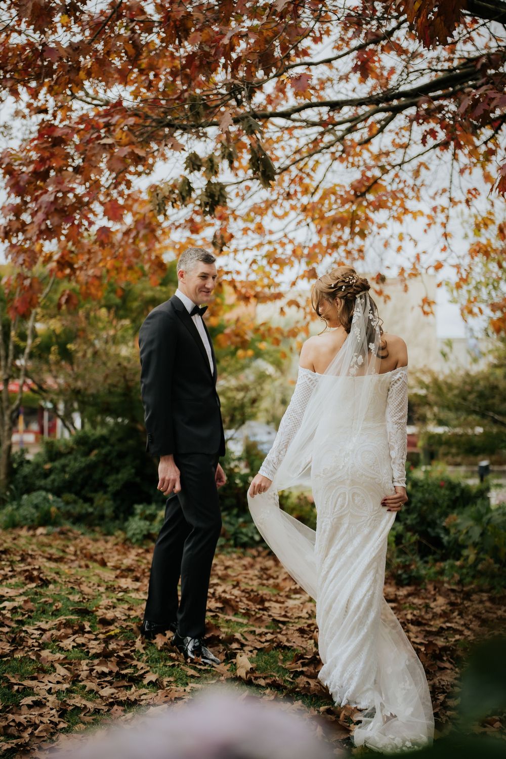 Bride and groom share a romantic moment under autumn foliage, she in lace gown with veil, he in black tuxedo.