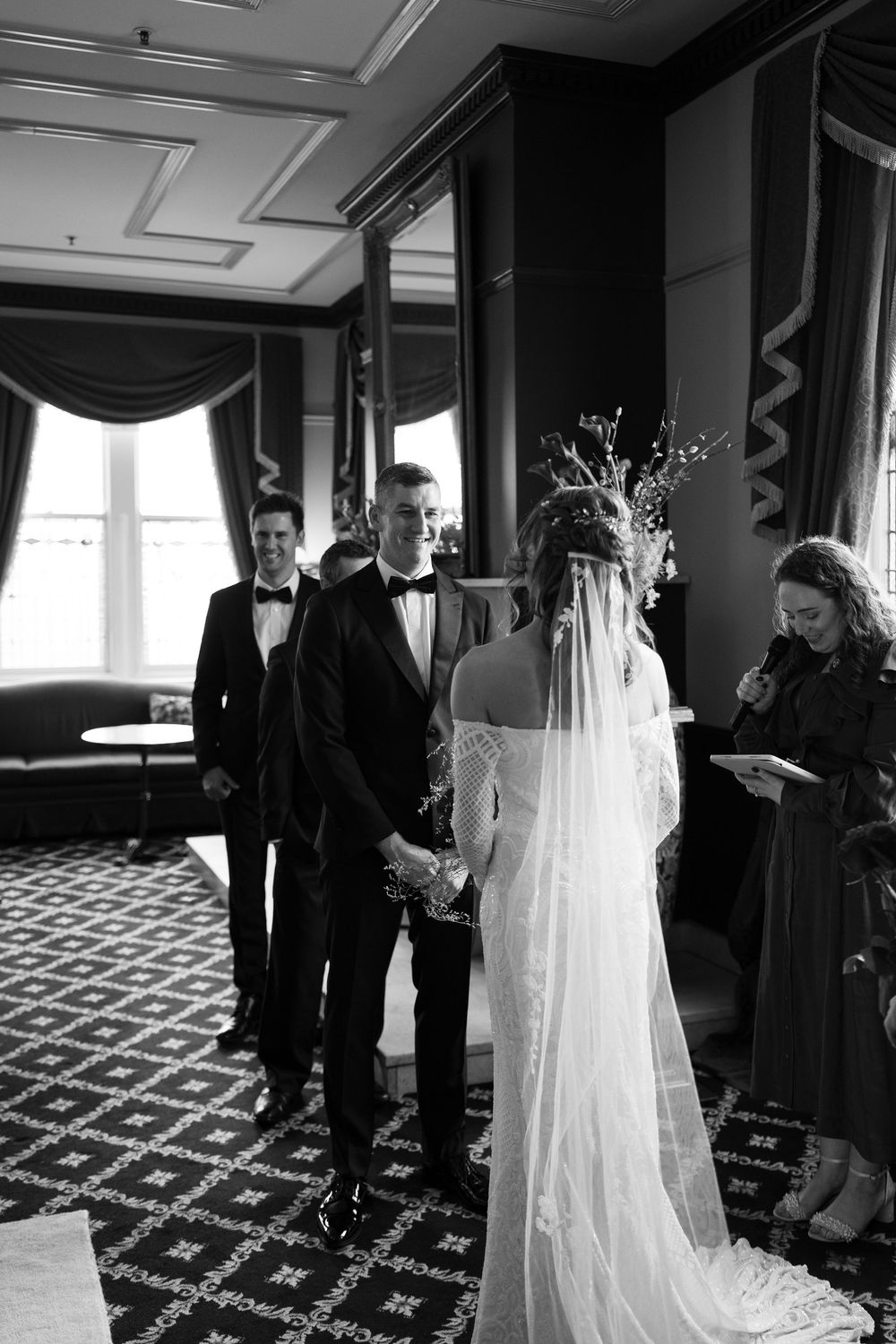 Black and white wedding ceremony photo of bride and groom holding hands at altar with officiant and guest in elegant venue.