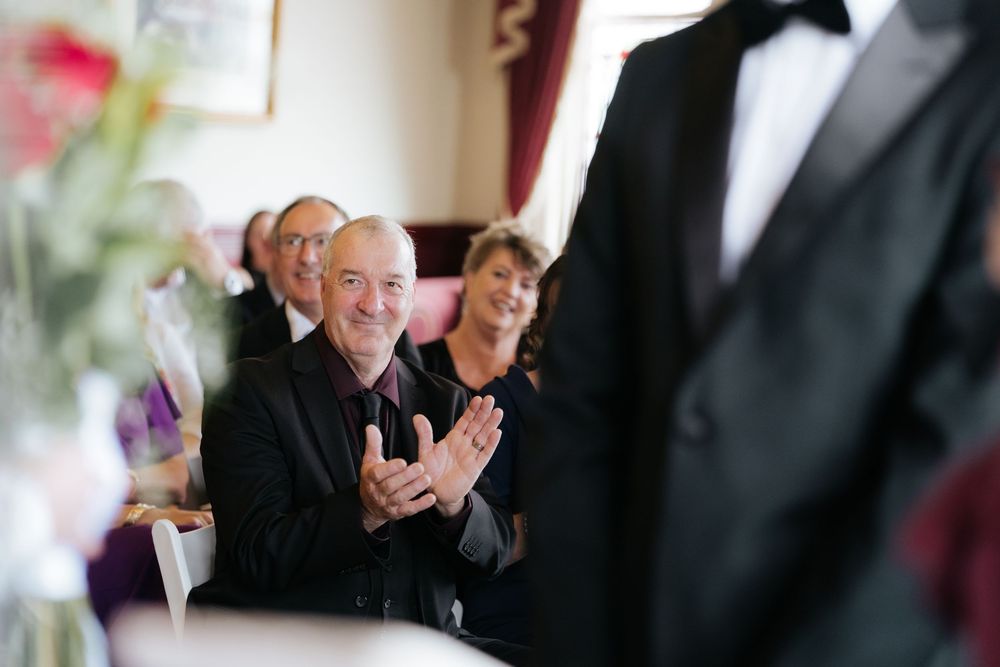 Smiling man in black suit clapping at a formal wedding reception event.