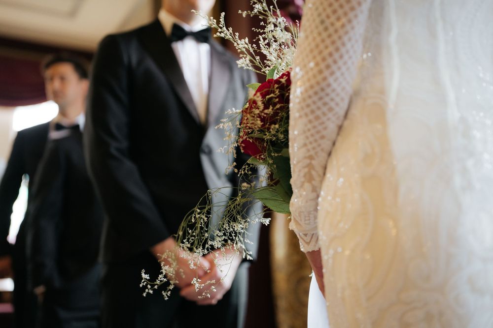 Bride and groom holding hands at wedding ceremony, bride holding red and white bouquet with lace dress.