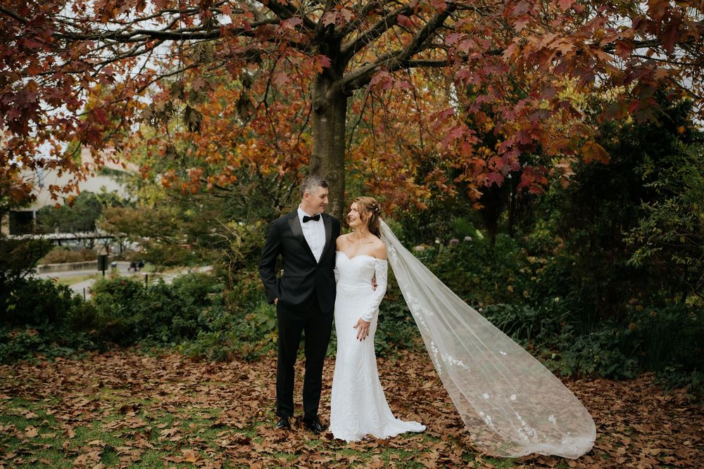 Bride and groom stand among autumn leaves beneath a colorful tree, bride wearing a flowing gown with cathedral veil.