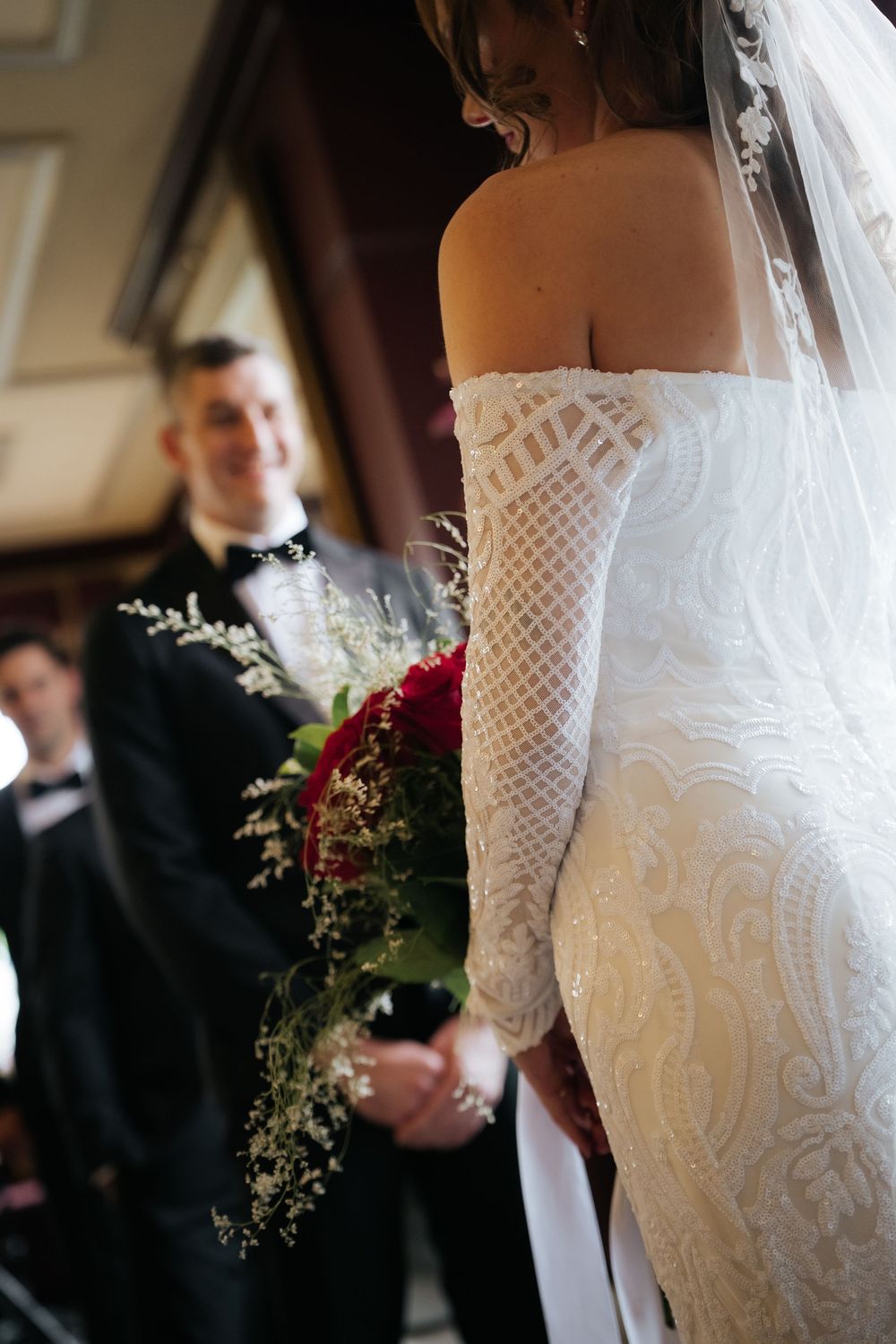 Bride in off-shoulder lace gown holds red bouquet facing smiling groom in black tuxedo at wedding ceremony.