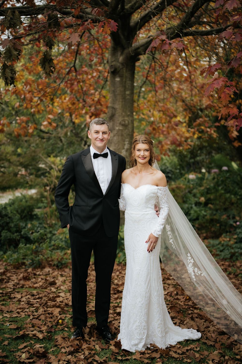 Bride and groom posing in autumn garden, she in lace off-shoulder gown with veil, he in black tuxedo.