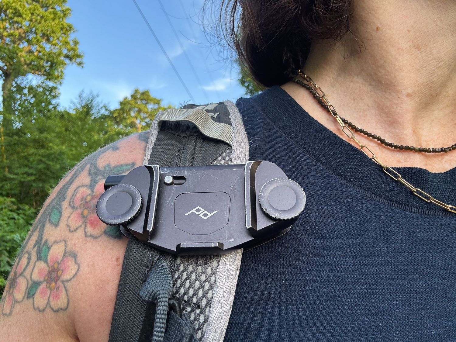 Person wearing a Peak Design camera clip on a backpack strap, with tattoos visible and trees in background.