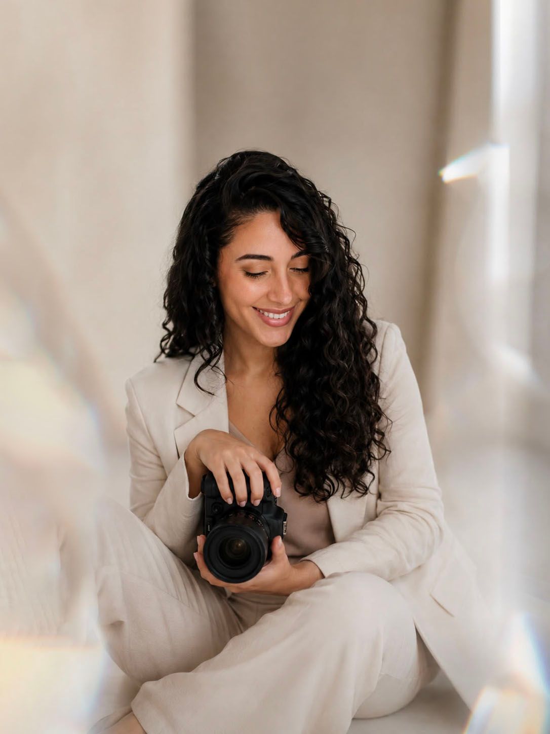 Black and white photo of a smiling young woman with curly hair wearing a hat and holding a camera.