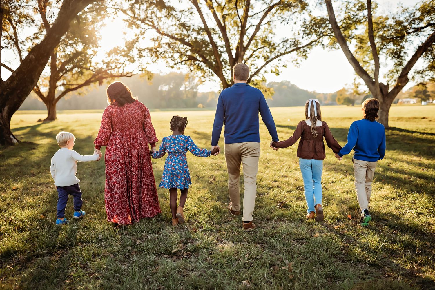 Family of five walking hand-in-hand through a sunlit park, viewed from behind on a warm autumn day.