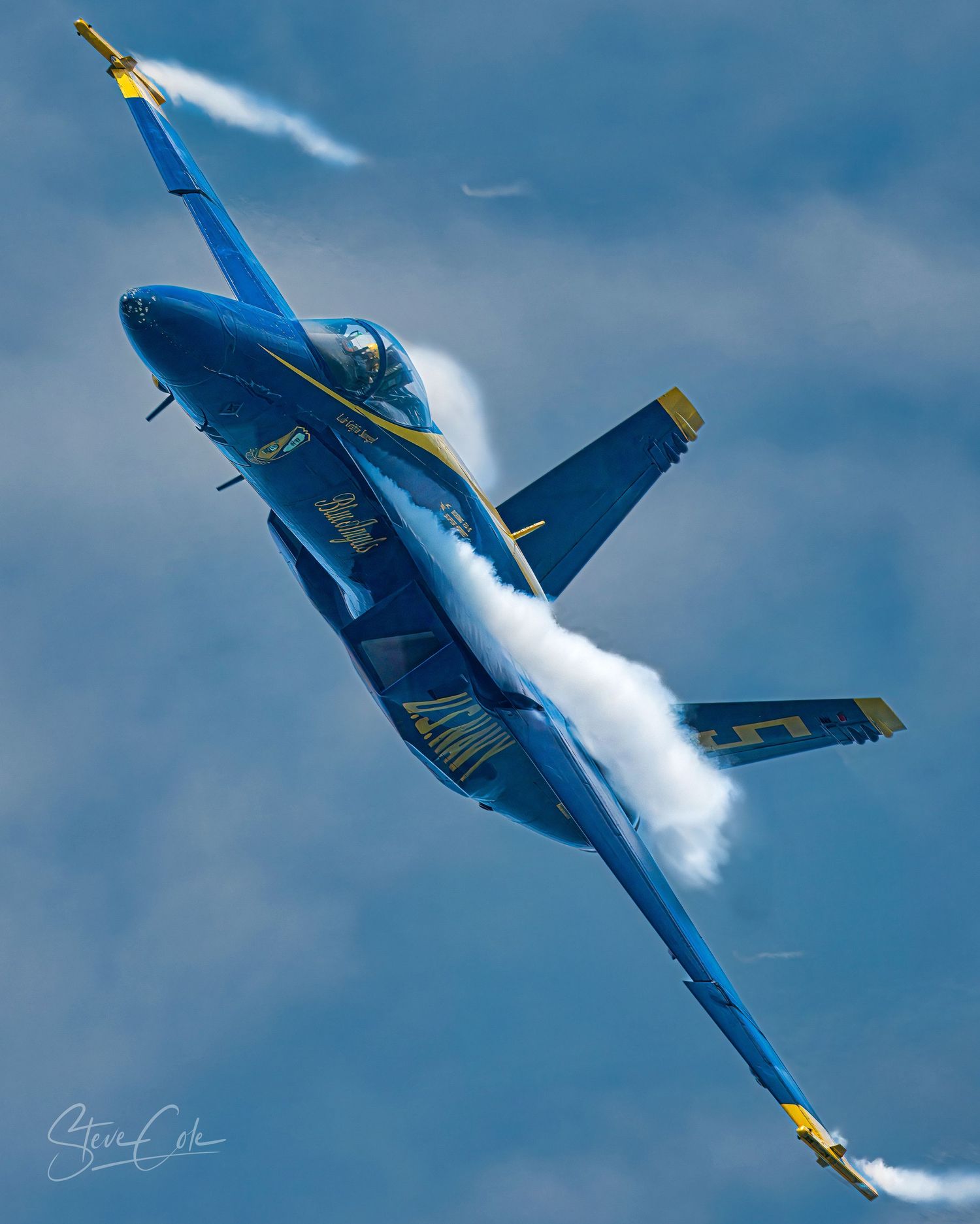 Blue Angels F/A-18 jet banked steeply with vapor trails forming over the wings against a blue sky, photo by Steve Cole Photography.