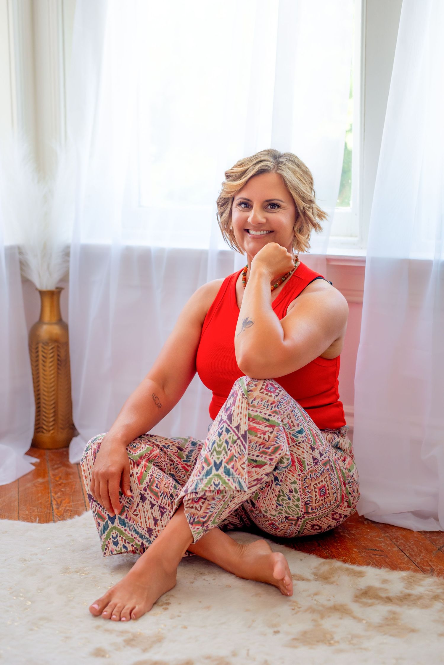Smiling blonde woman sitting cross-legged on a rug in a red top and colorful patterned pants near a bright window.