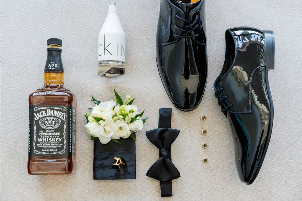 Groom's wedding flat lay with Jack Daniel's whiskey, Calvin Klein cologne, black patent leather shoes, bow tie, and white flowers.