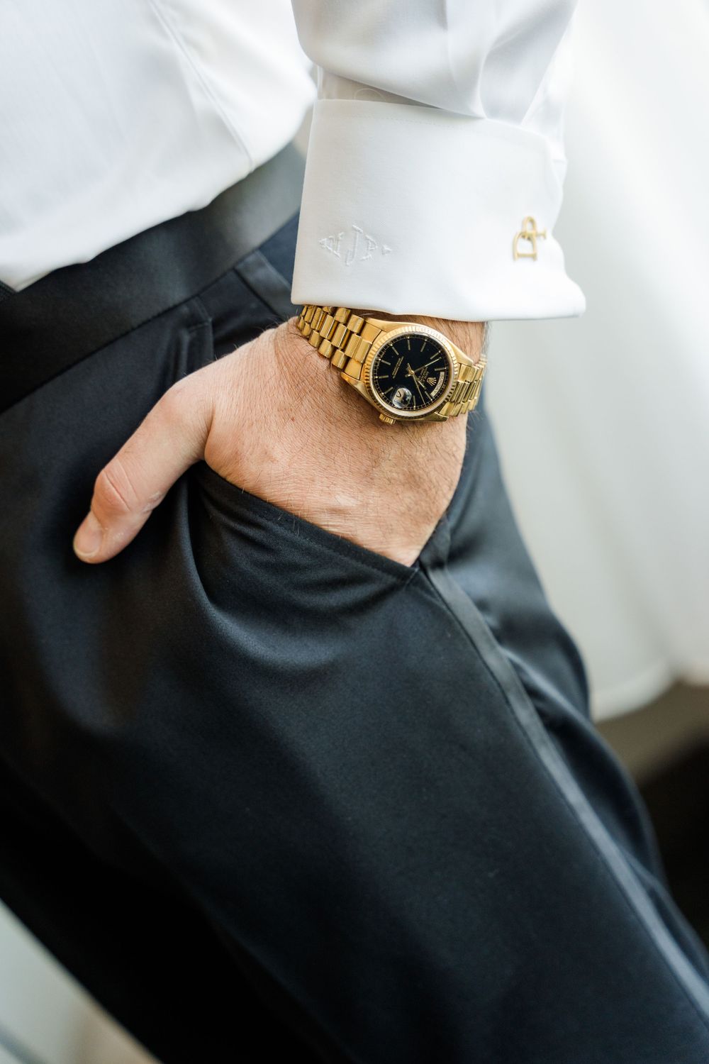 Man in white dress shirt with gold cufflinks and luxury gold watch with black dial, hand in dark trouser pocket.