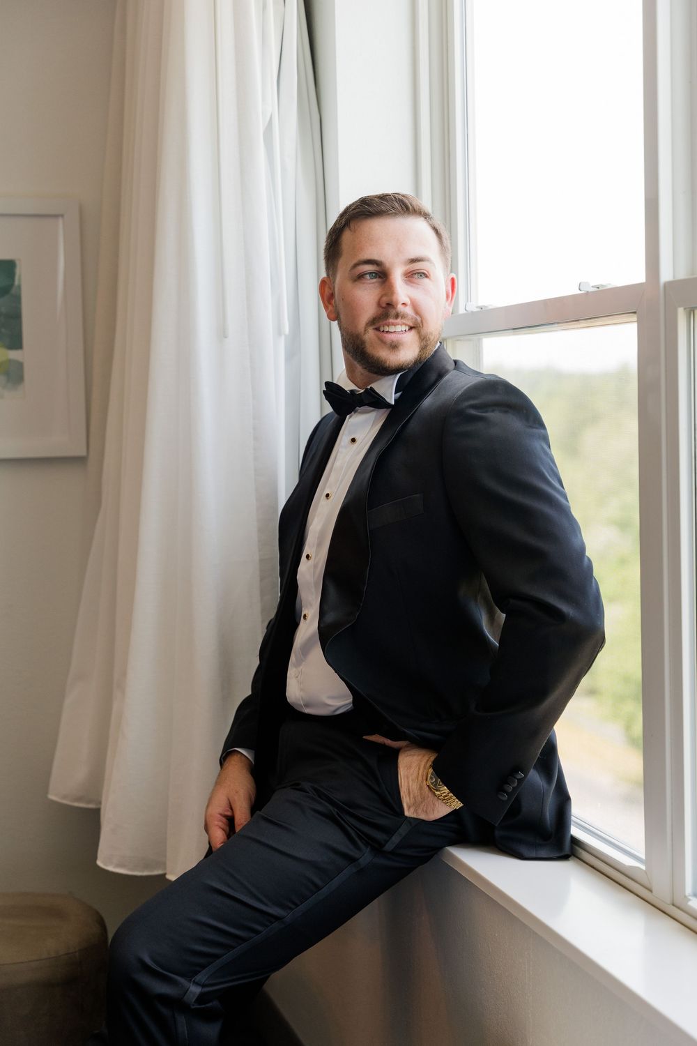 A dapper groom in a black tuxedo with bow tie sits on a window sill, smiling confidently in a bright room.