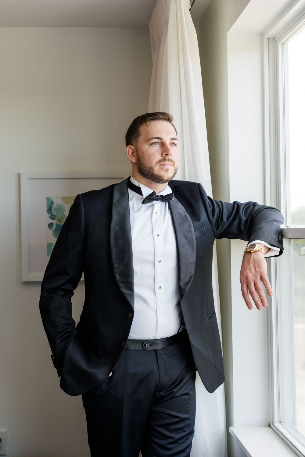Groom in black tuxedo with bow tie and gold watch leaning against window, looking outside on wedding day.