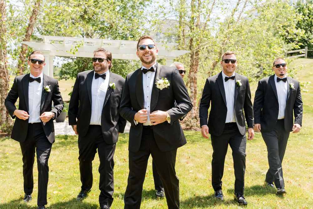 Groom and groomsmen in black tuxedos and bow ties with sunglasses walking outdoors at a wedding.