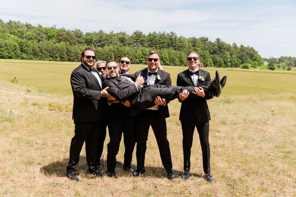 Groomsmen in black suits and sunglasses lifting the groom outdoors in a sunny green field on his wedding day.