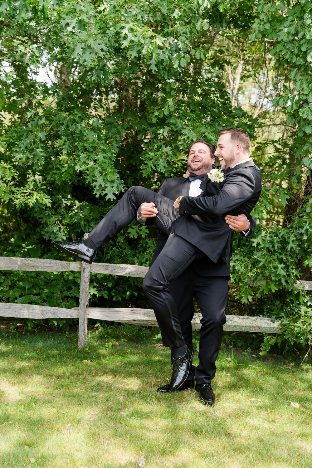 Two grooms in black tuxedos laugh as one carries the other outdoors near a wooden fence.