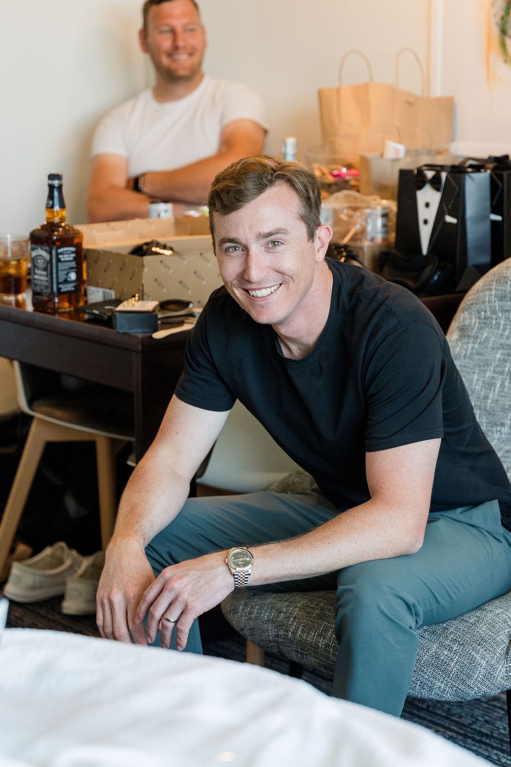 Smiling man in black t-shirt sitting on bed at groomsmen party with Jack Daniels whiskey and gift bags.