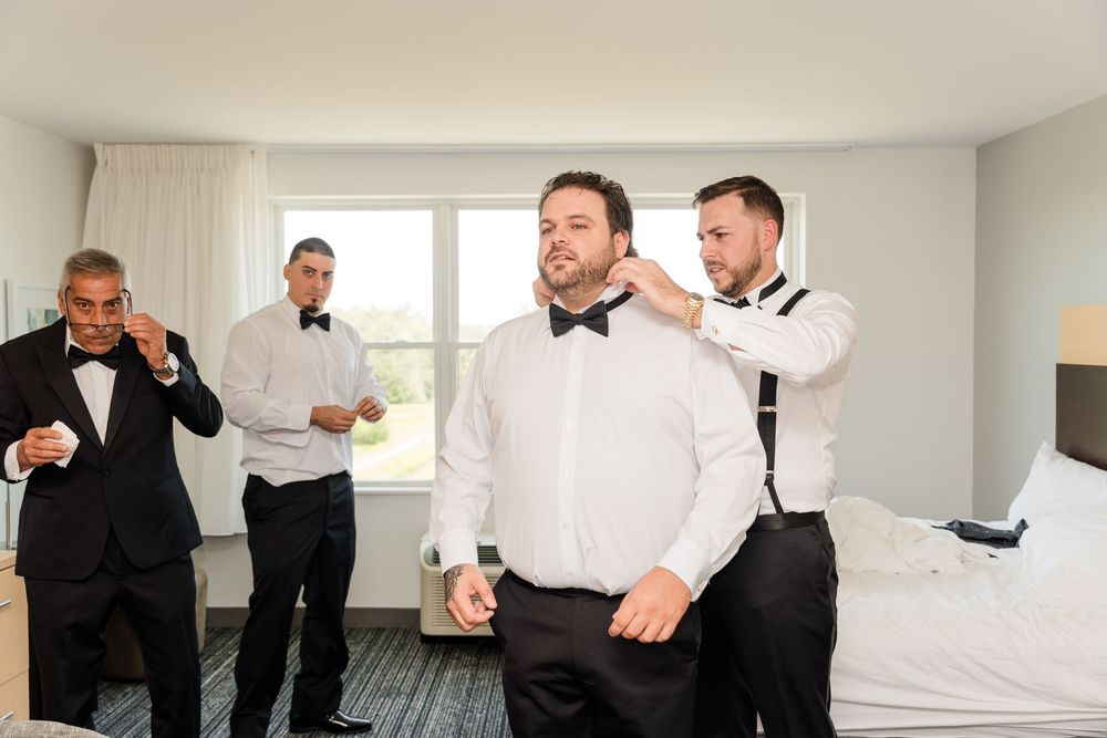 Groomsmen helping groom adjust bow tie in hotel room before wedding ceremony.