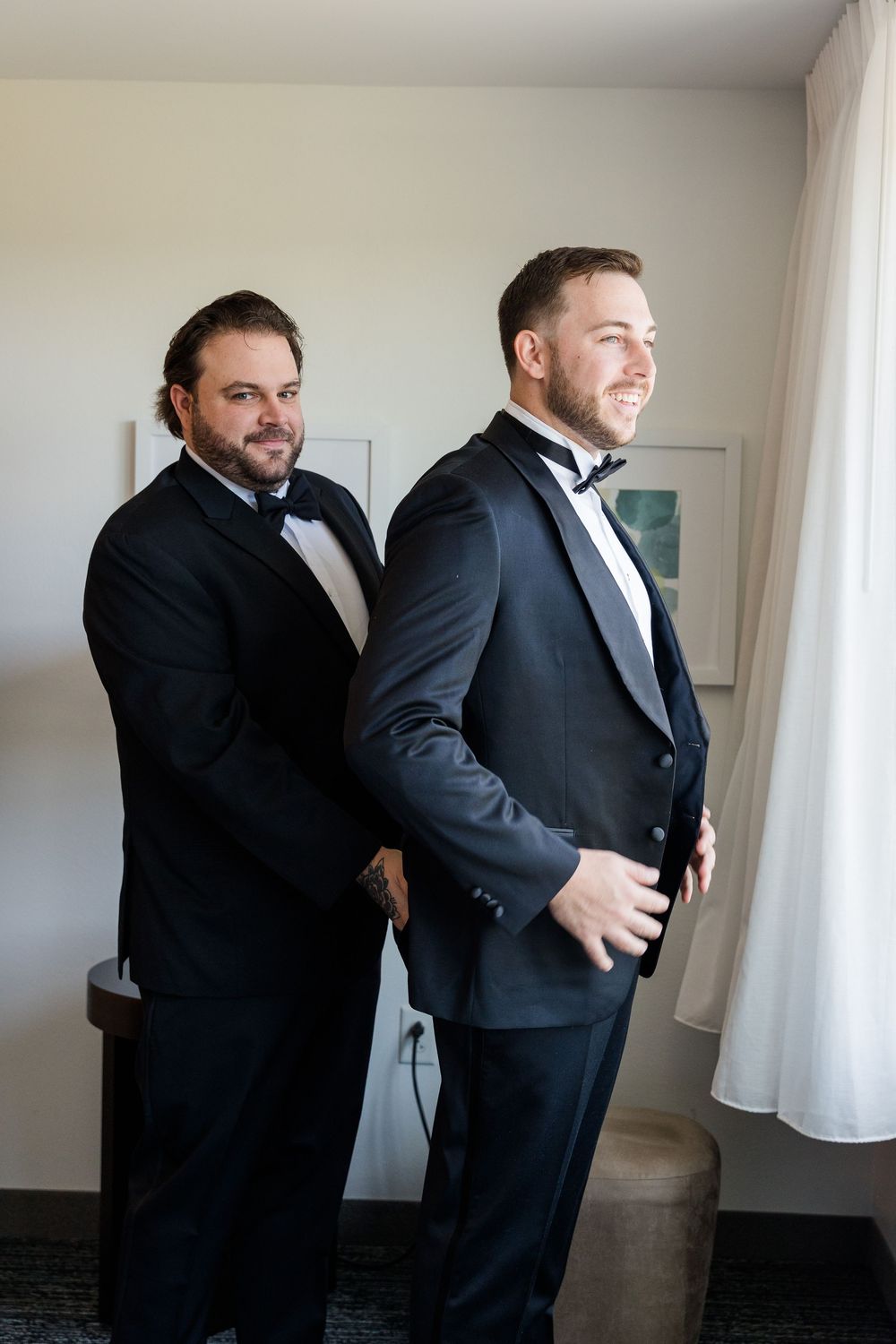 Two men in black tuxedos with bow ties getting ready, one helping the other button his jacket.
