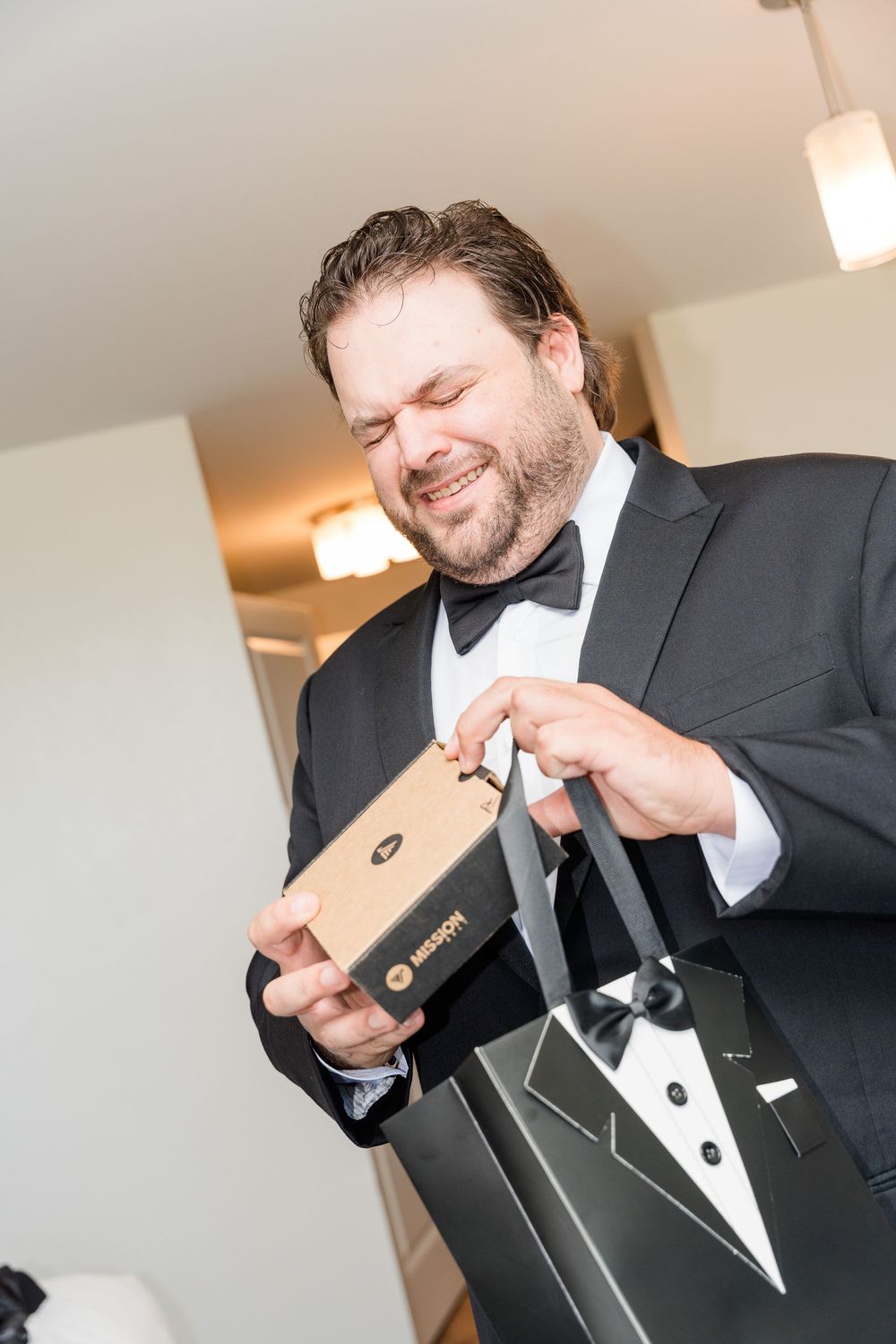Smiling groom in tuxedo opening a gift box at his wedding, holding a tuxedo-themed gift bag.