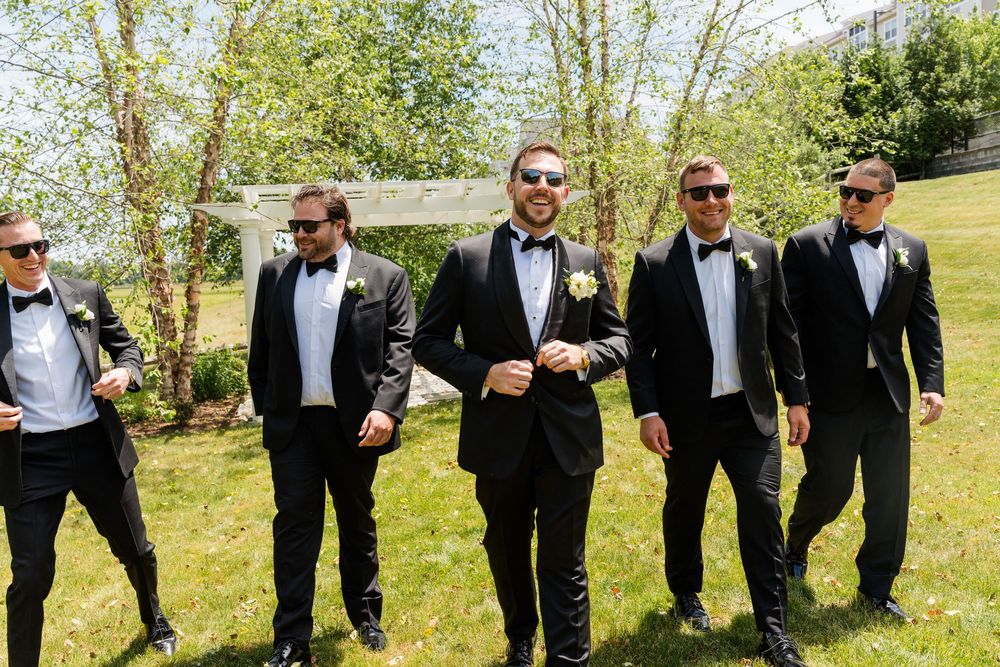 Groom and groomsmen in black tuxedos with bow ties and boutonnieres walking outdoors on a sunny wedding day.