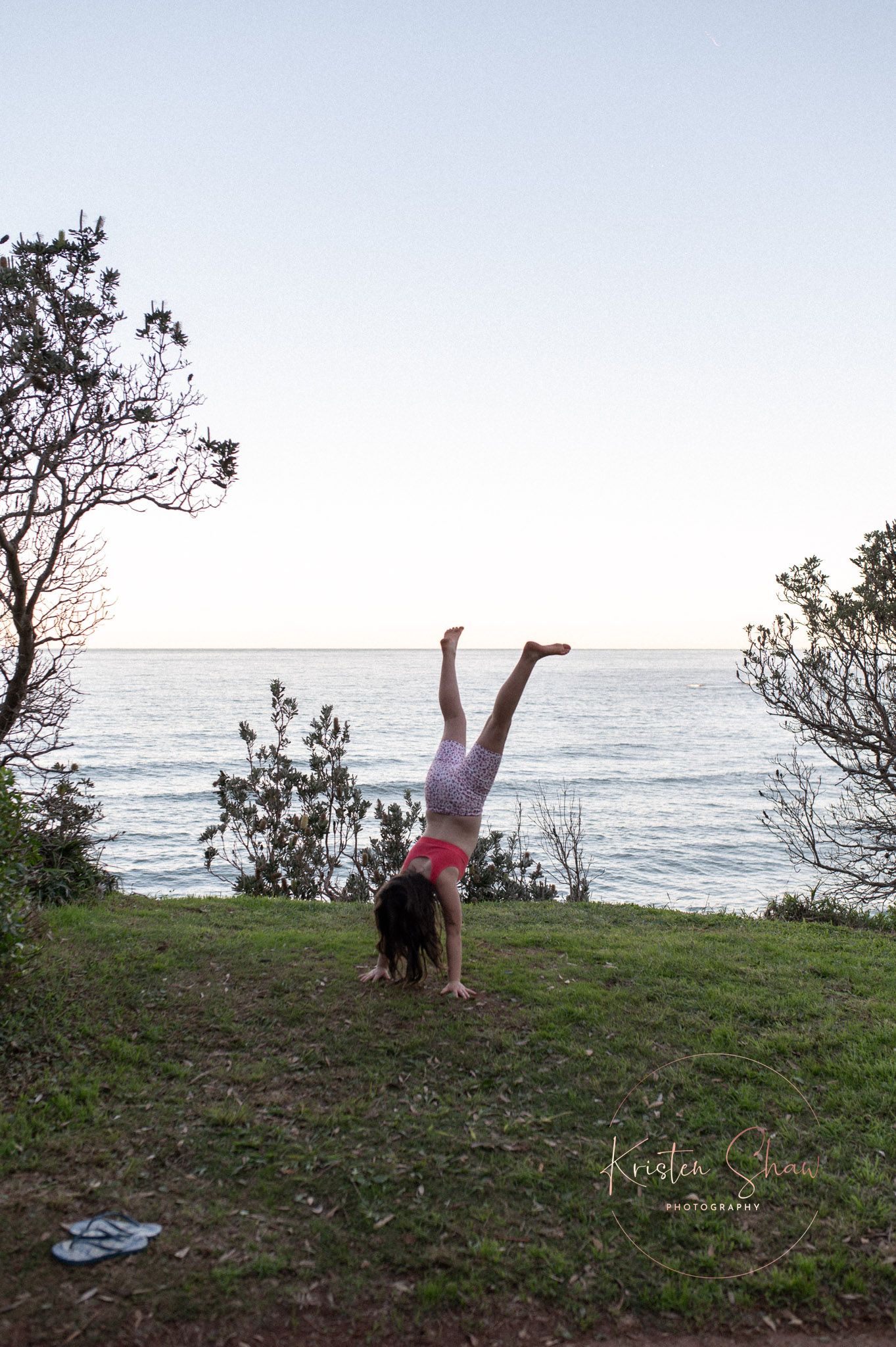 Woman doing a handstand on grass overlooking the ocean at dusk near trees.