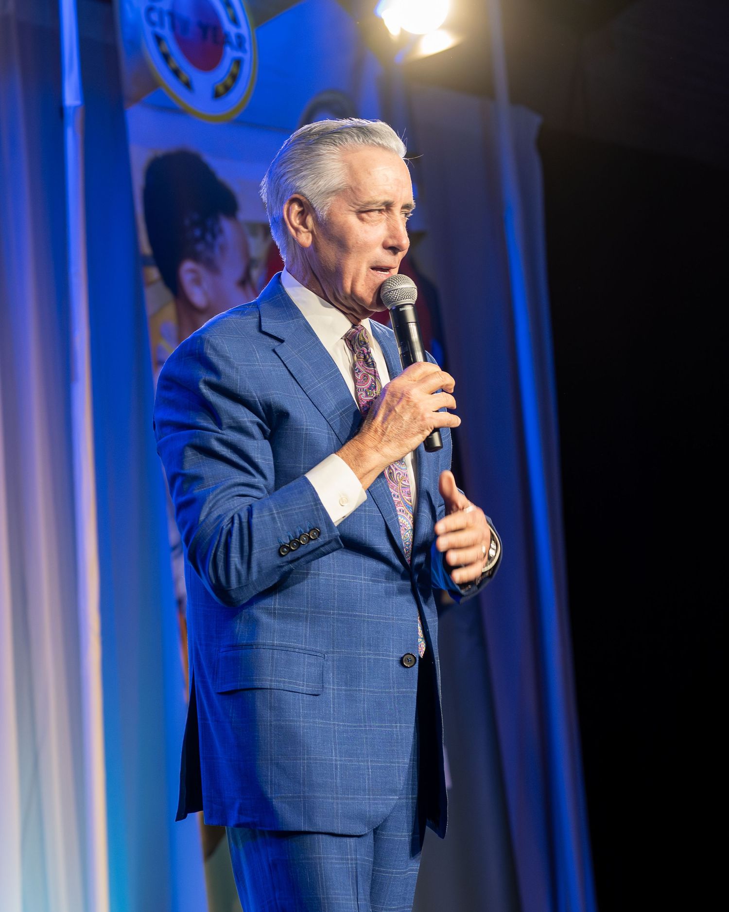 Man in blue suit speaking into microphone on stage at formal event  fundraising gala with spotlight.