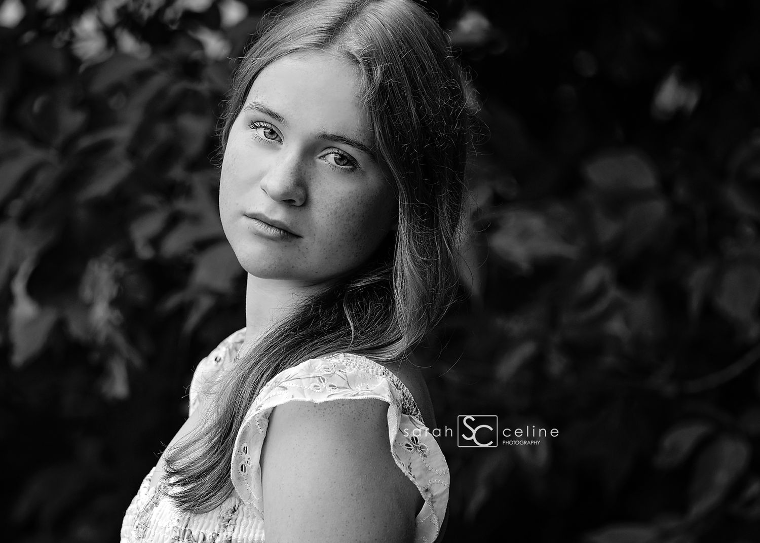 Black and white portrait of a young woman with long hair posing outdoors among lush foliage.