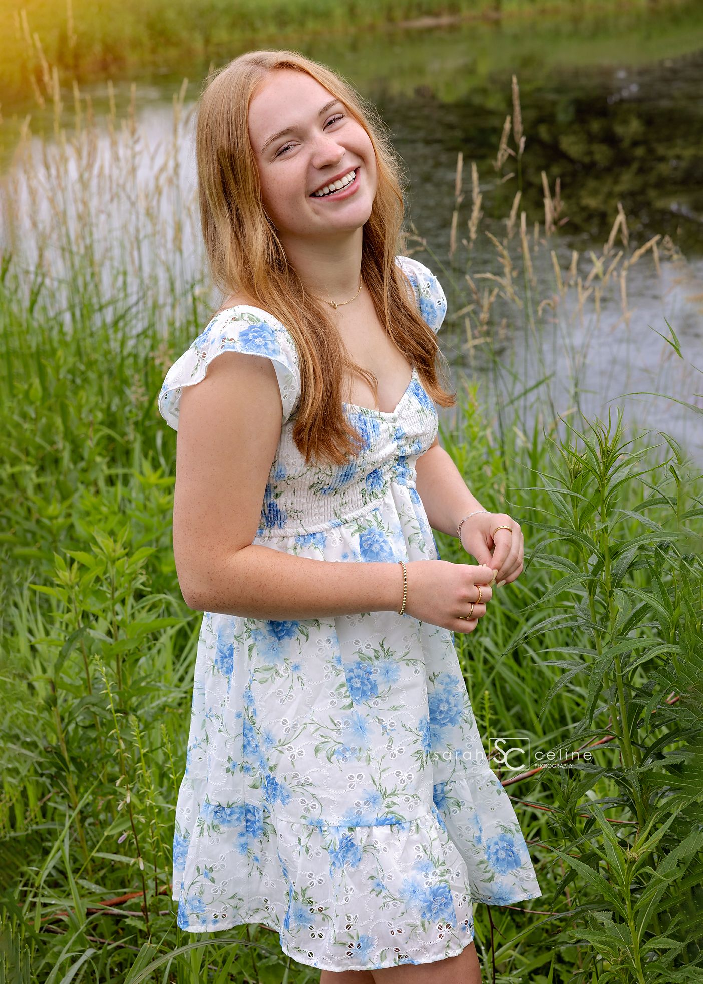 Smiling teenage girl in a white floral dress standing in a lush green field near a pond on a sunny day.