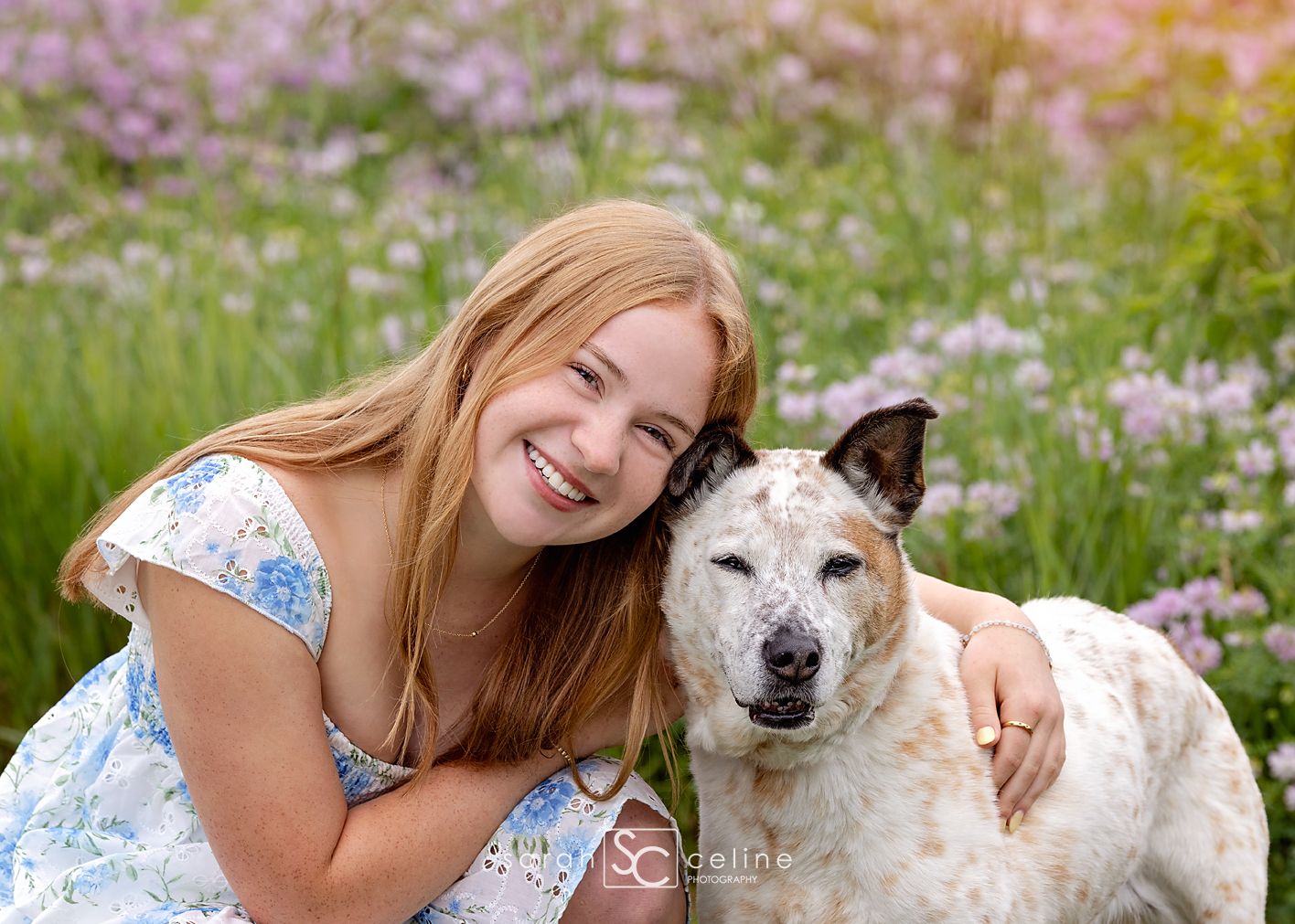 Smiling young woman with long red hair hugging her dog in a colorful wildflower field.
