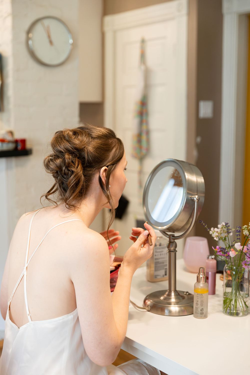 Bride in white dress applying makeup at vanity mirror with elegant updo hairstyle while getting ready for wedding day in London, Ontario.