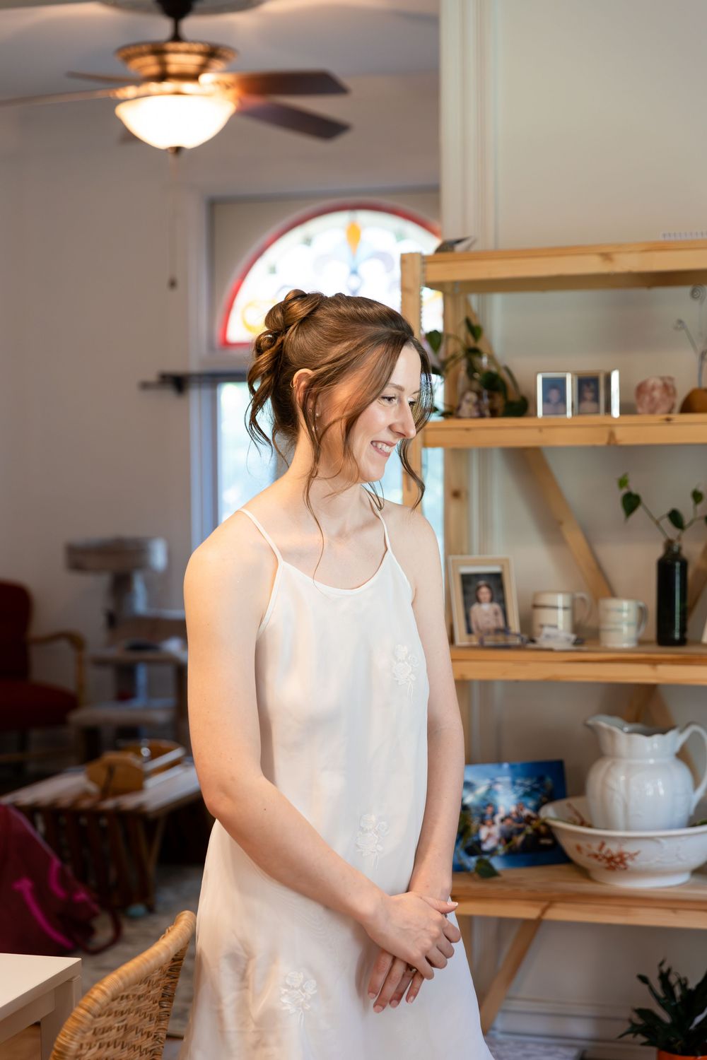 Smiling bride in white slip dress stands near wooden shelves in a warmly decorated room with ceiling fan in London, Ontario.