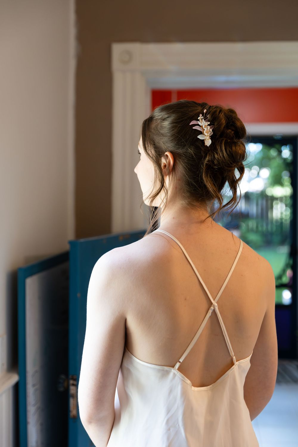 Bride stands with her back towards the camera looking out a window in a white nightgown on the morning of her london, ontario wedding.