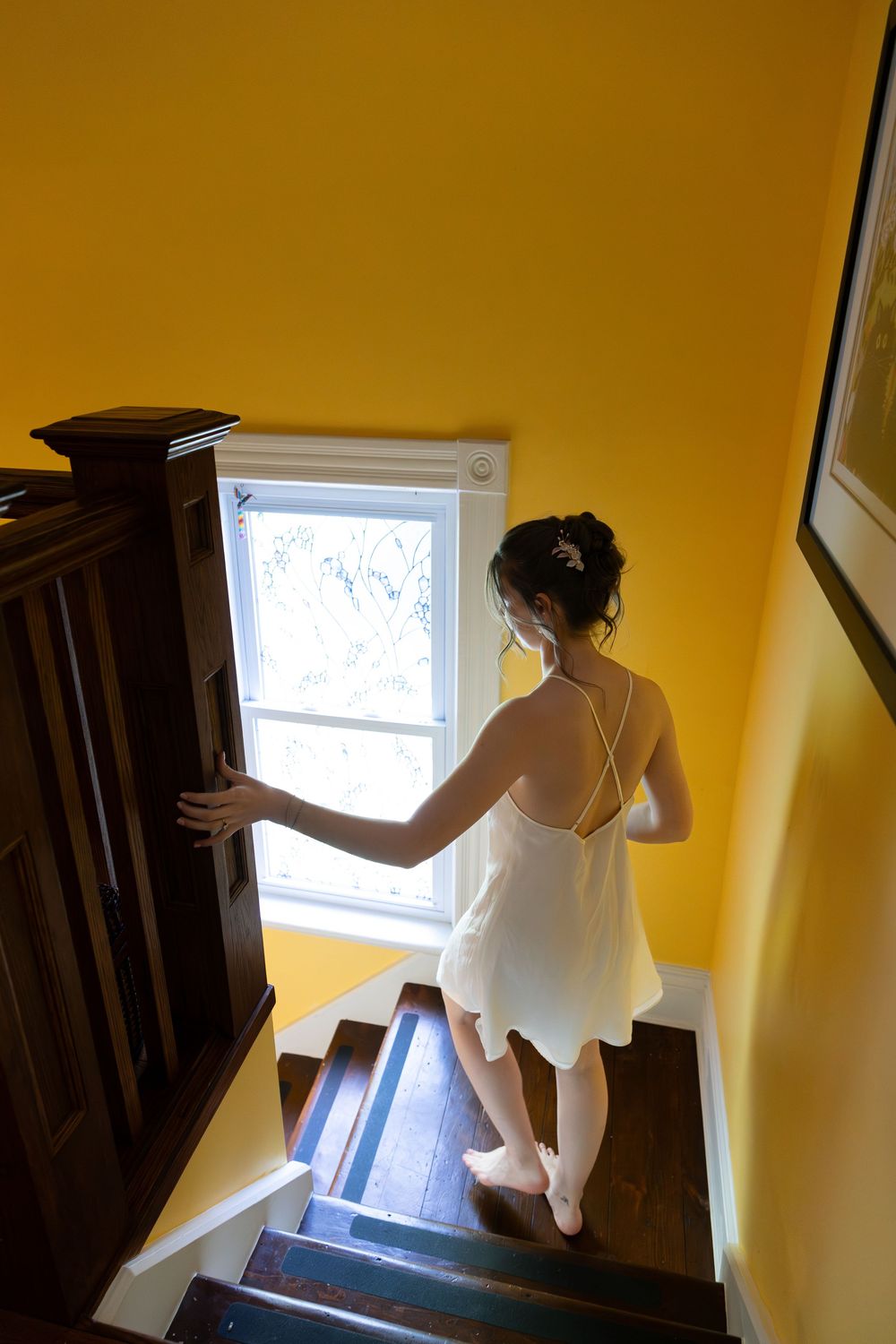 Bride in white dress descending wooden stairs near a bright window in a yellow-walled interior on the morning of a london, ontario wedding.