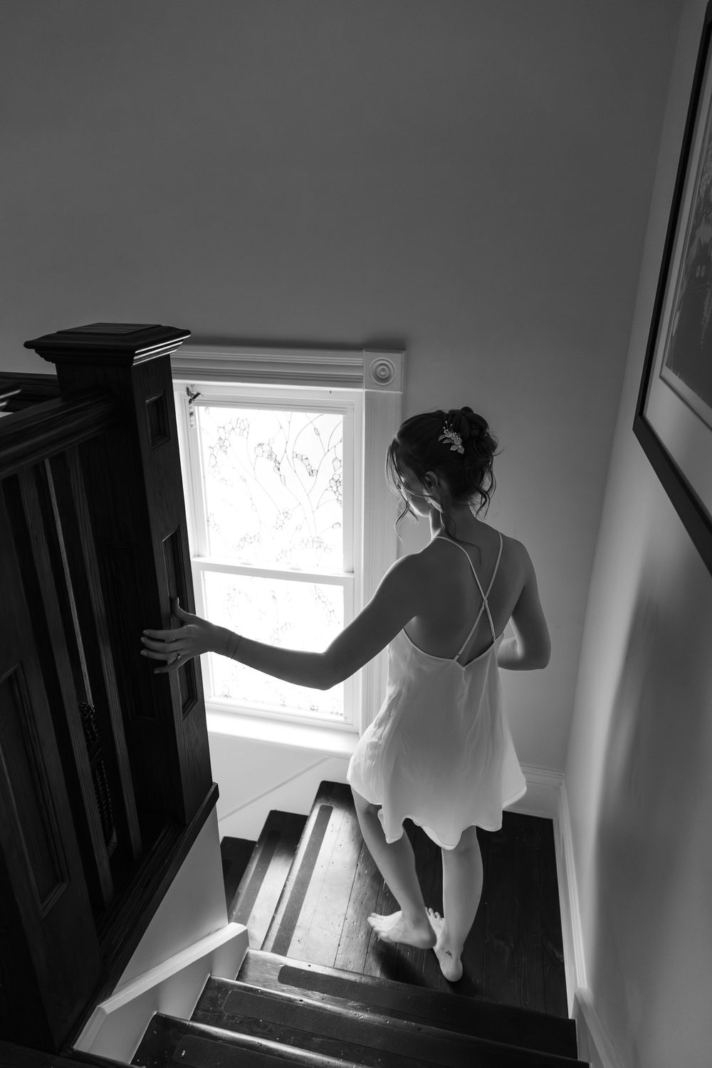 Black and white photo of bride in a white slip dress descending stairs toward a bright window on the morning of a london, ontario weddding.