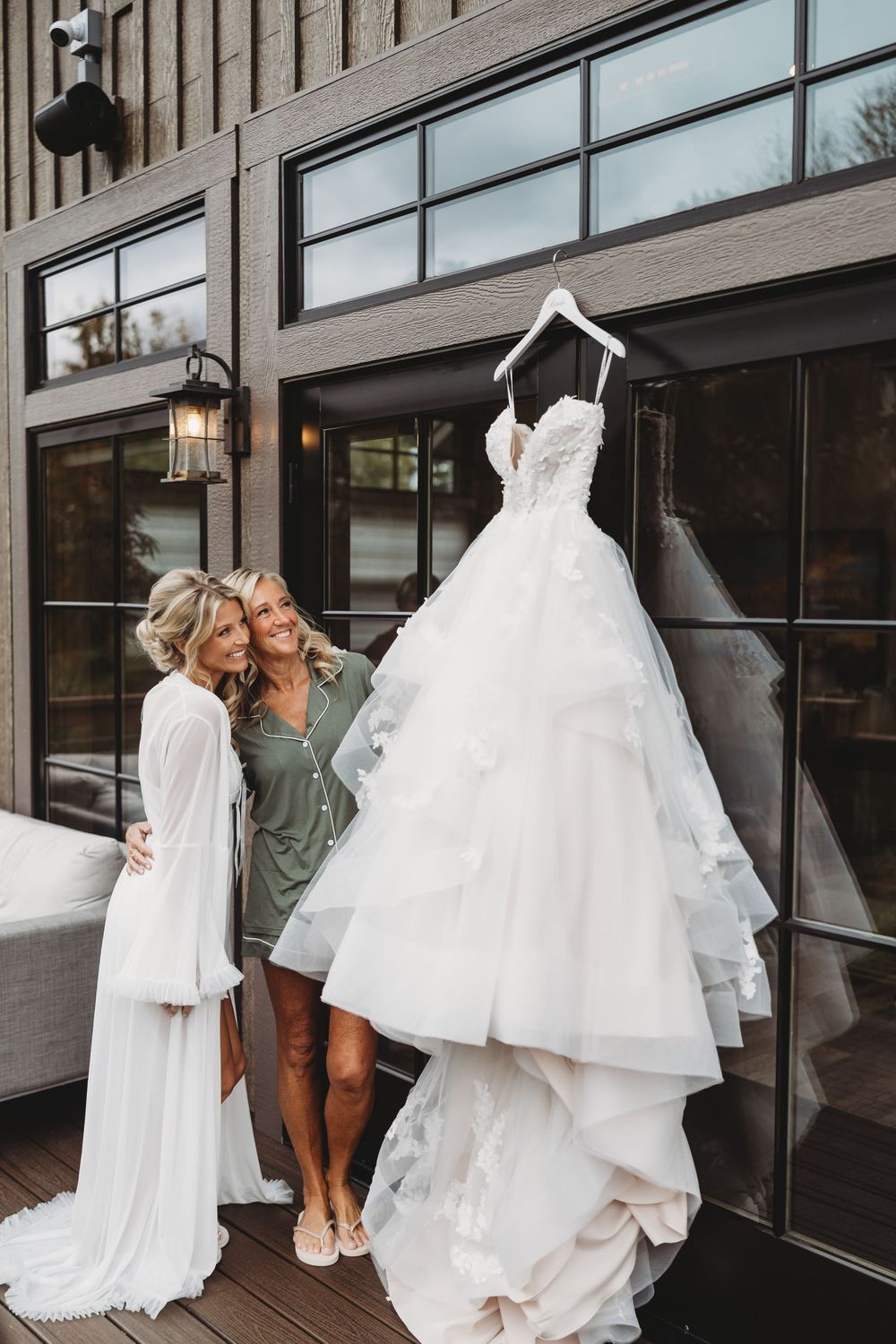 Bride in white robe admires her layered tulle wedding gown hanging outside a modern venue with a friend.