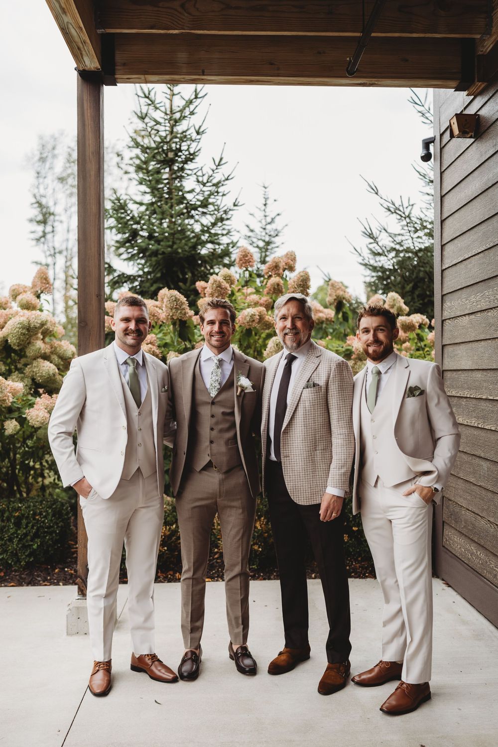 Four groomsmen in tan and beige suits with brown shoes pose outside a wedding venue surrounded by hydrangeas.