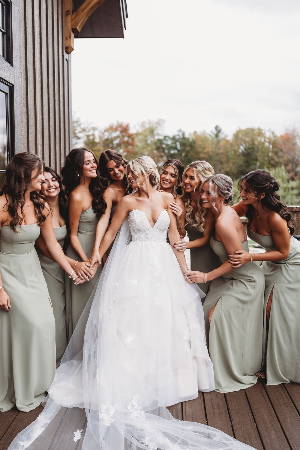 Bride in strapless lace ballgown surrounded by smiling bridesmaids in sage green dresses on wooden deck.