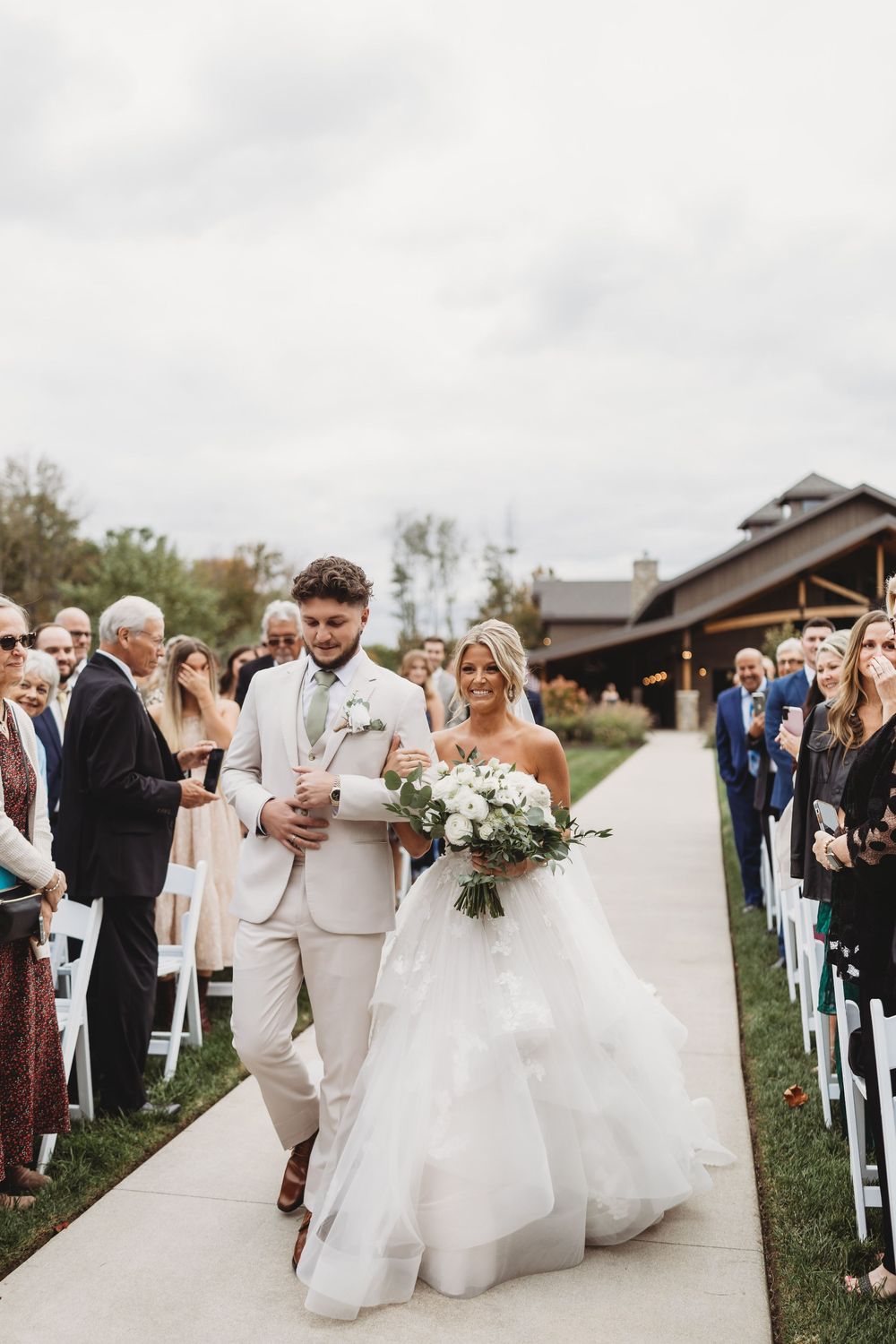 Smiling bride and escort walk down outdoor wedding aisle as guests look on.