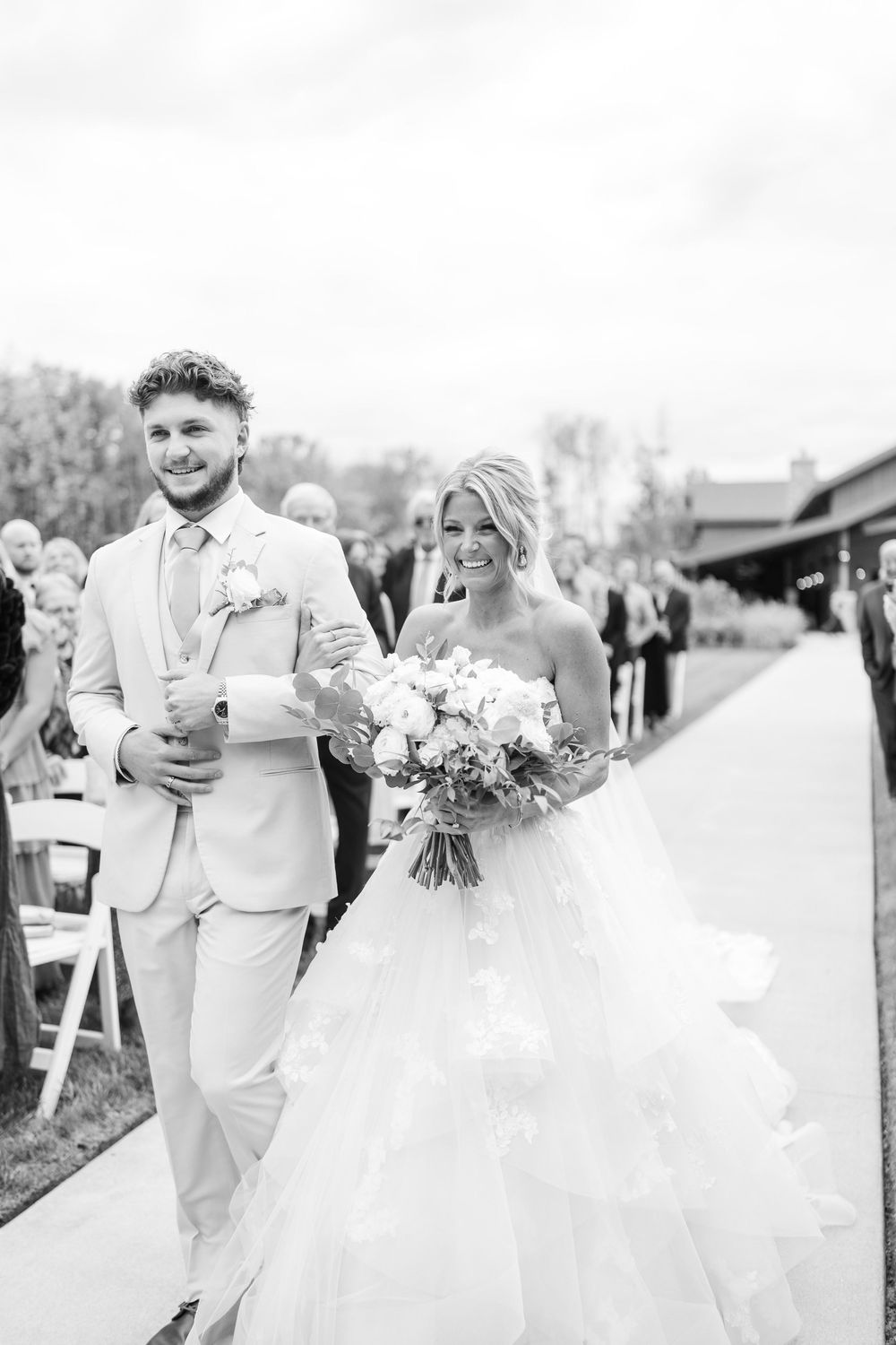 Black and white photo of beaming bride and escort walking down wedding ceremony aisle.