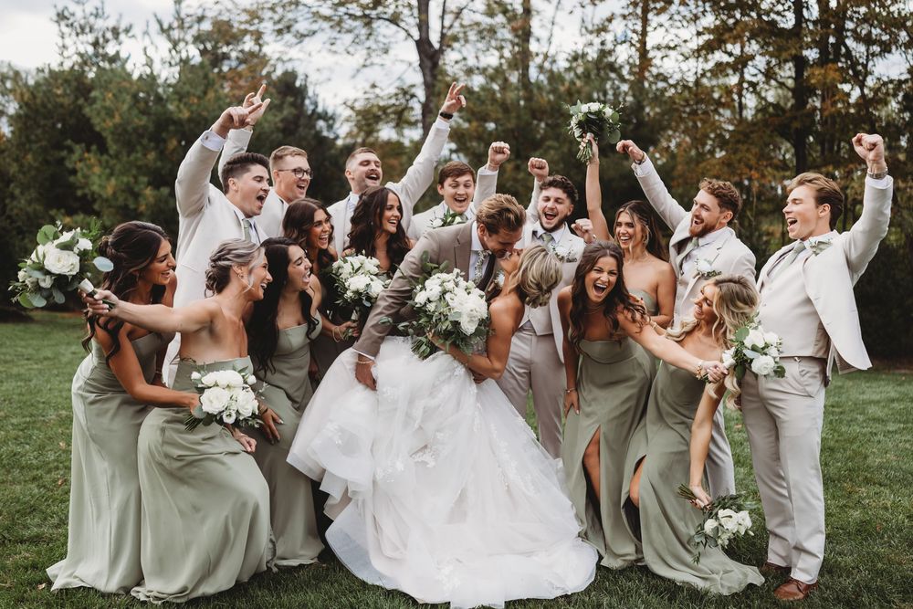 Joyful wedding party celebrating outdoors with bride and groom kissing, bridesmaids in sage green gowns, groomsmen in white suits.