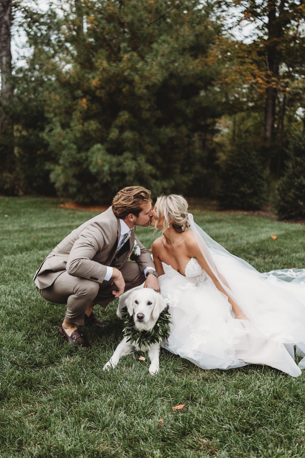 Bride and groom kissing over their white golden retriever wearing floral wreath on green lawn.