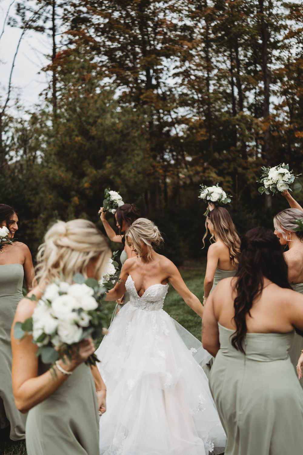 Bride in white lace gown walks with bridesmaids in sage green dresses, holding white floral bouquets outdoors.