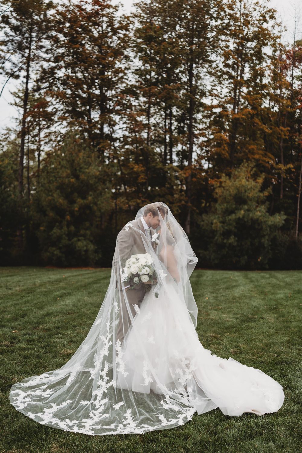 Bride and groom kissing under flowing lace cathedral veil on green lawn with colorful autumn trees.