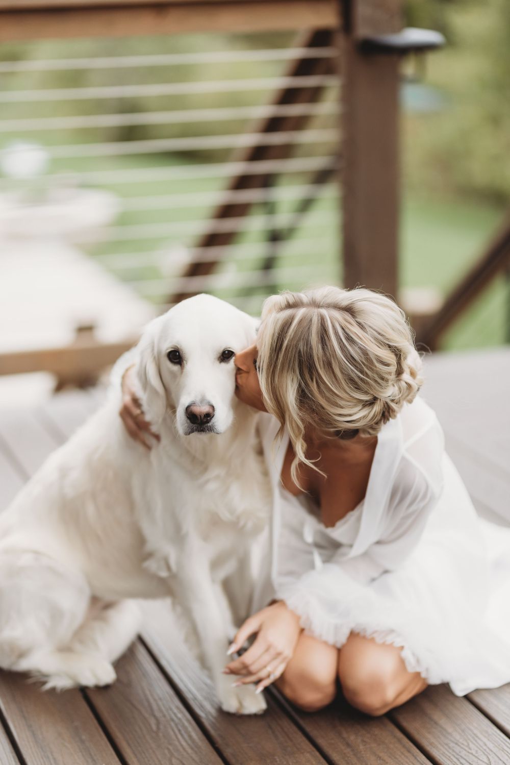 Bride in white robe kissing white golden retriever dog on wooden deck during wedding morning prep.