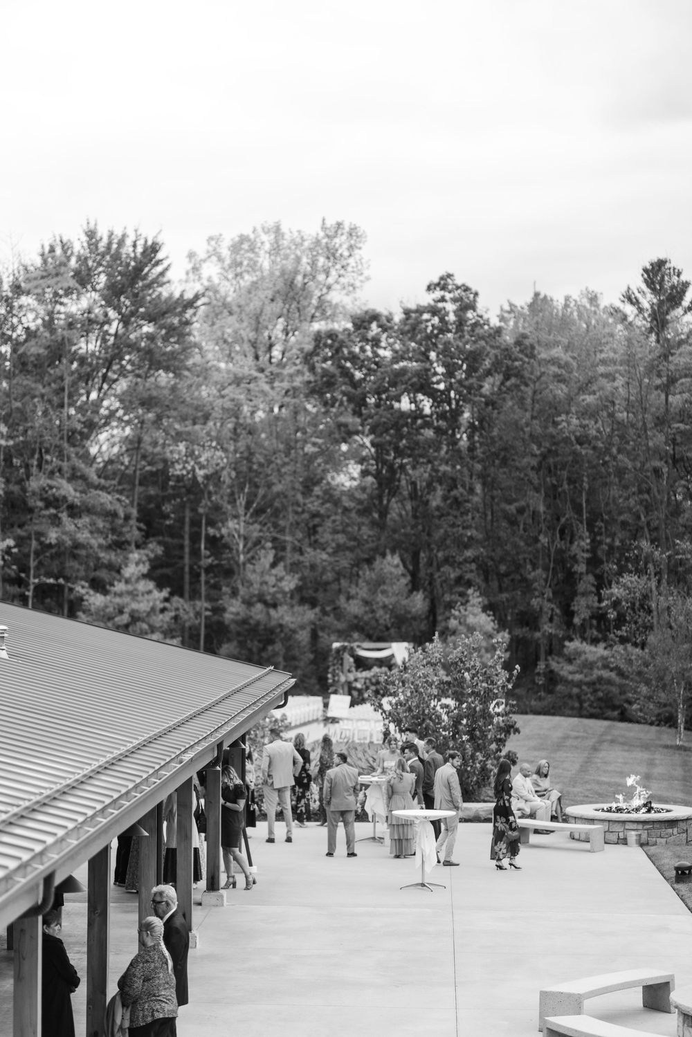 Black and white aerial view of wedding guests mingling on outdoor patio near forest.