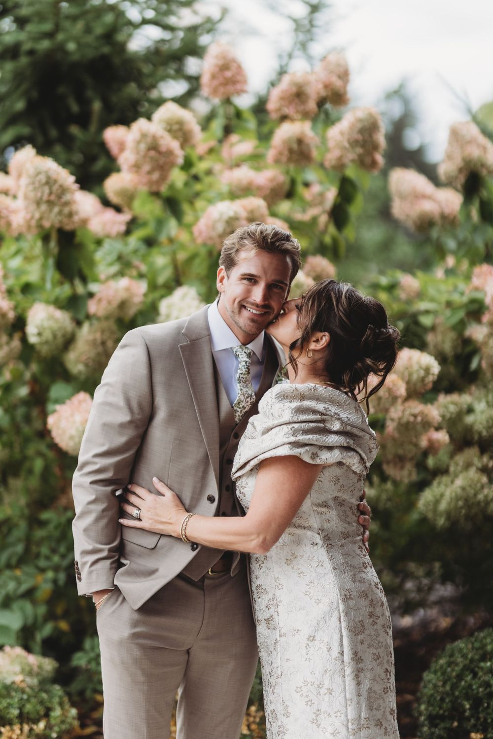 Smiling groom in tan suit embraced by bride in lace gown before blooming hydrangea bushes in garden setting.