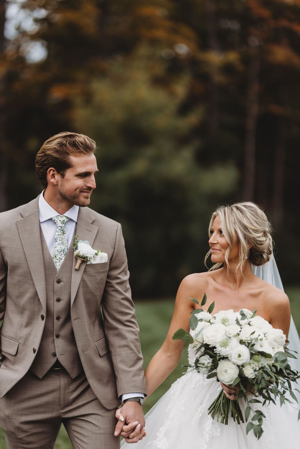 Bride and groom holding hands walking outdoors in fall, groom in tan suit, bride in white gown holding white floral bouquet.